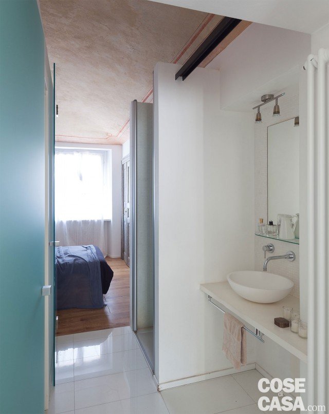 Due porte a battente in vetro satinato chiudono, il blocco centrale dell’antibagno-bagno, evidenziato da un inserto a terra in marmo ricomposto lucido che interrompe il parquet. L’illuminazione d’ambiente è prodotta da tubi al neon nascosti da barre in ferro fissate a parete. Due porte a battente in vetro satinato chiudono, il blocco centrale dell’antibagno-bagno, evidenziato da un inserto a terra in marmo ricomposto lucido che interrompe il parquet. L’illuminazione d’ambiente è prodotta da tubi al neon nascosti da barre in ferro fissate a parete.