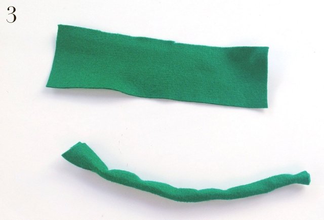 Ritagliare un rettangolo di stoffa verde di circa 20x6 cm, avvolgerlo su se stesso dalla parte lunga, fino ad ottenere un tubicino. Ritagliare un rettangolo di stoffa verde di circa 20x6 cm, avvolgerlo su se stesso dalla parte lunga, fino ad ottenere un tubicino.