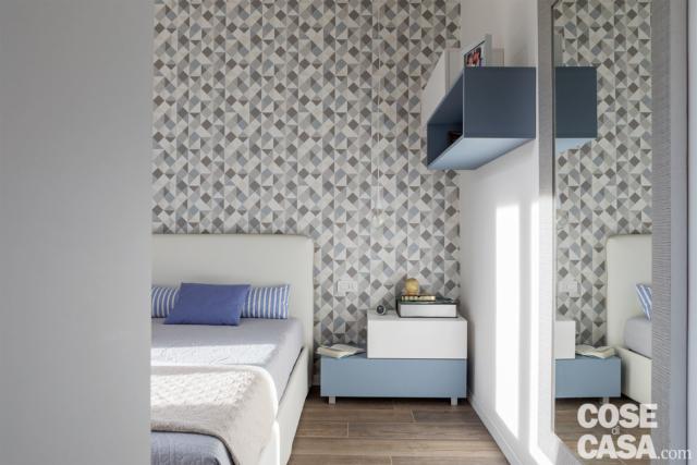 camera matrimoniale con letto tessile, parete di fondo rivestita in carata da parati a motivo geometrico sui toni del grigio. comodino in laccato bianco e azzurro, pavimento in gres effetto legno camera matrimoniale con letto tessile, parete di fondo rivestita in carata da parati a motivo geometrico sui toni del grigio. comodino in laccato bianco e azzurro, pavimento in gres effetto legno