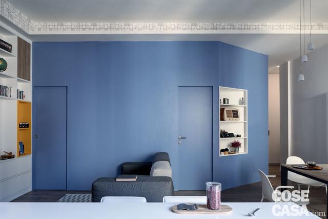 In soggiorno, parete smaltata in azzurro con le porte del bagno e dello studio rifinite nello stesso colore, divano, mobile a giorno, tavolo da pranzo. In primo piano penisola della cucina In soggiorno, parete smaltata in azzurro con le porte del bagno e dello studio rifinite nello stesso colore, divano, mobile a giorno, tavolo da pranzo. In primo piano penisola della cucina