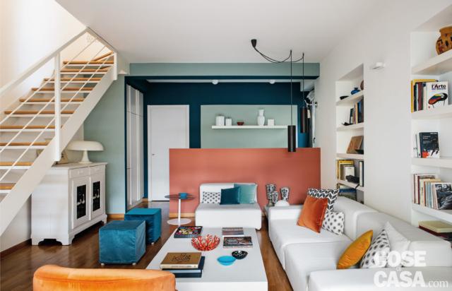 Zona conversazione con divano bianco e poltrona, pouf turchesi e muretto divisorio color corallo, librerie, scala Zona conversazione con divano bianco e poltrona, pouf turchesi e muretto divisorio color corallo, librerie, scala
