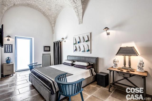 La camera matrimoniale è attigua al soggiorno, con cui condivide i segni architettonici distintivi del piano “nobile” della palazzina: volte di pietra Roccato di Ostuni lasciate a vista e pavimentazione di chianche di pietra Bronzetto di Trani. Anche qui pochi arredi di carattere declinati in tinte neutre, per rendere protagonisti l’involucro costruttivo e la luce naturale, che entra copiosa dalla portafinestra modello Prolux Evolution di Oknoplast. Il letto Poncio di Ethos, dai dettagli sartoriali, è sovrastato da un’opera d’arte tridimensionale e materica, così come materici sono i comodini a fianco, di Marchetti Maison. Completano l’insieme un tavolo-console di artigianato locale e una piccola cassettiera Moab 80, oltre alle due poltroncine InOut 724 di Gervasoni laccate in una originale sfumatura di blu industriale. L’illuminazione è risolta con apparecchi a parete Ambra di Anna Lari uguali a quelli della zona giorno, per dare uniformità stilistica al piano. La camera matrimoniale è attigua al soggiorno, con cui condivide i segni architettonici distintivi del piano “nobile” della palazzina: volte di pietra Roccato di Ostuni lasciate a vista e pavimentazione di chianche di pietra Bronzetto di Trani. Anche qui pochi arredi di carattere declinati in tinte neutre, per rendere protagonisti l’involucro costruttivo e la luce naturale, che entra copiosa dalla portafinestra modello Prolux Evolution di Oknoplast. Il letto Poncio di Ethos, dai dettagli sartoriali, è sovrastato da un’opera d’arte tridimensionale e materica, così come materici sono i comodini a fianco, di Marchetti Maison. Completano l’insieme un tavolo-console di artigianato locale e una piccola cassettiera Moab 80, oltre alle due poltroncine InOut 724 di Gervasoni laccate in una originale sfumatura di blu industriale. L’illuminazione è risolta con apparecchi a parete Ambra di Anna Lari uguali a quelli della zona giorno, per dare uniformità stilistica al piano.