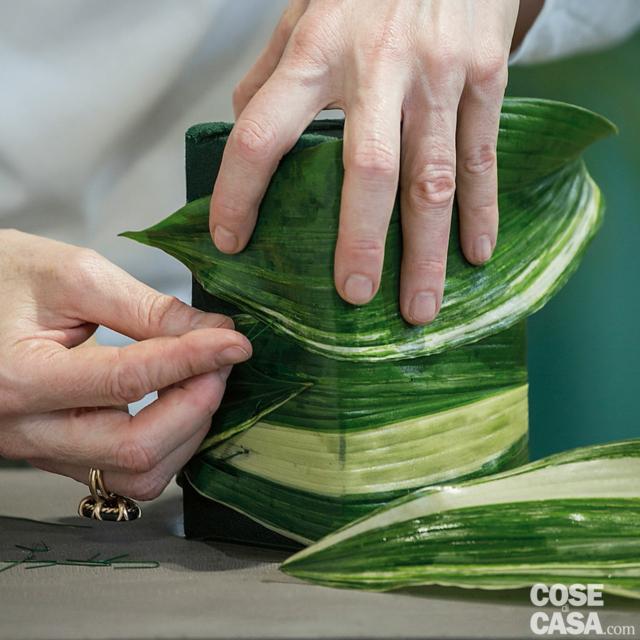 2. Con il coltellino eliminare gli steli delle foglie di Aspidistra, quindi pulire le lamine da polvere e residui di terra. Tagliare il filo di ferro con il tronchesino per ottenere tanti segmenti di circa 4 cm, che piegati, formeranno delle graffette. Avvolgere la prima foglia intorno alla spugna e fissarla a ogni giro con le graffette, quindi ripetere con la seconda foglia; proseguire fino a rivestire completamente la spugna. 2. Con il coltellino eliminare gli steli delle foglie di Aspidistra, quindi pulire le lamine da polvere e residui di terra. Tagliare il filo di ferro con il tronchesino per ottenere tanti segmenti di circa 4 cm, che piegati, formeranno delle graffette. Avvolgere la prima foglia intorno alla spugna e fissarla a ogni giro con le graffette, quindi ripetere con la seconda foglia; proseguire fino a rivestire completamente la spugna.