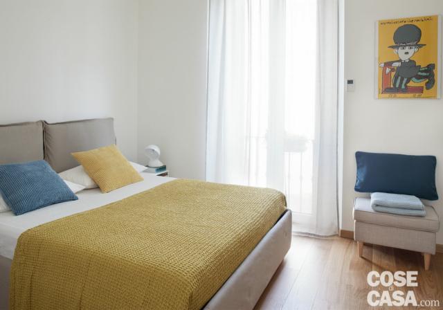 bilocale, camera matrimoniale, letto tessile, copriletto giallo, cuscini giallo e azzurro, portafinestra, pouf, pavimento in parquet bilocale, camera matrimoniale, letto tessile, copriletto giallo, cuscini giallo e azzurro, portafinestra, pouf, pavimento in parquet