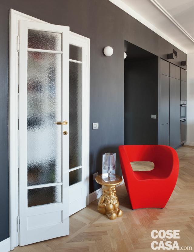 bilocale aperto, porta in legno laccato a doppio battente, vetro serigrafato, parete in finitura scura, applique sferica, poltroncina rossa e tavolino di design, parquet a spina di pesce bilocale aperto, porta in legno laccato a doppio battente, vetro serigrafato, parete in finitura scura, applique sferica, poltroncina rossa e tavolino di design, parquet a spina di pesce