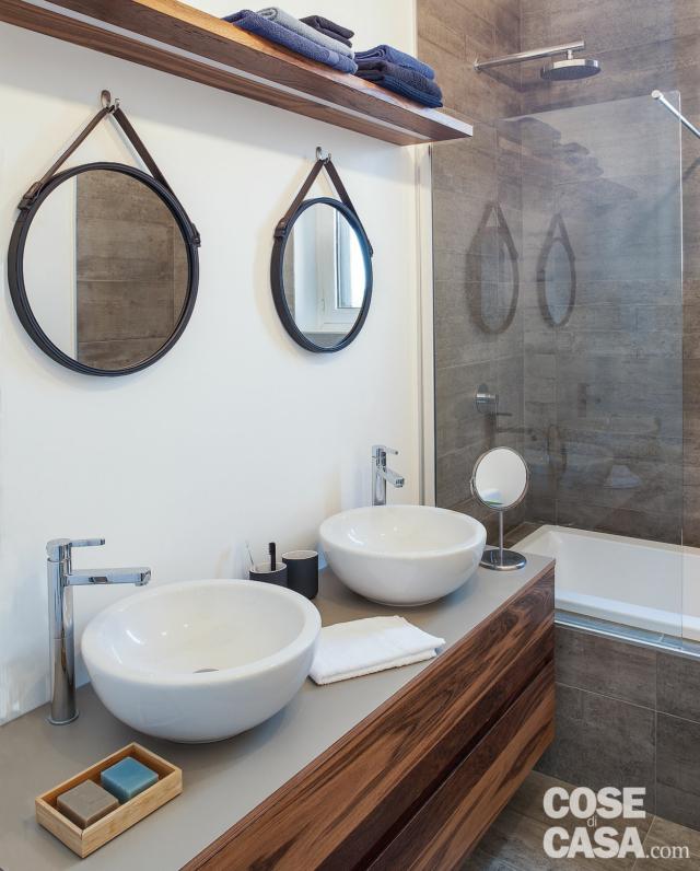 bagno, piano con due lavabi, rubinetti a colonna, specchi tondi, ripiano, vasca, divisorio vetrato, rivestimento in gres grigio bagno, piano con due lavabi, rubinetti a colonna, specchi tondi, ripiano, vasca, divisorio vetrato, rivestimento in gres grigio