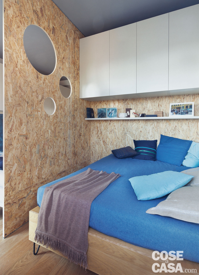 zona notte del monolocale, parete in OSB, pensili in laccato bianco, ripiano, letto, copriletto e cuscini azzurri zona notte del monolocale, parete in OSB, pensili in laccato bianco, ripiano, letto, copriletto e cuscini azzurri