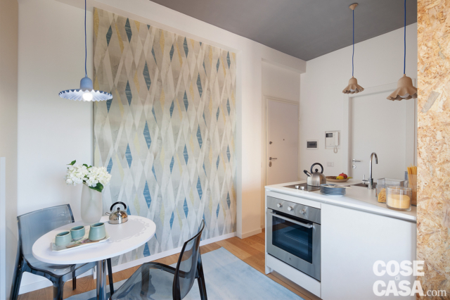 angolo cottura del monolocale con forno e piastra a induzione, lavello, lampade a sospensione in cartone sagomato, pannello in carta da parati, mini zona pranzo per due con tavolo rotondo, soffitto grigio angolo cottura del monolocale con forno e piastra a induzione, lavello, lampade a sospensione in cartone sagomato, pannello in carta da parati, mini zona pranzo per due con tavolo rotondo, soffitto grigio