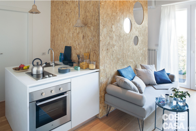 tramezzo angolare in OSB, blocco cucina con forno, piano cottura a induzione e lavello, divano grigio, coffee table e portafinestra tramezzo angolare in OSB, blocco cucina con forno, piano cottura a induzione e lavello, divano grigio, coffee table e portafinestra