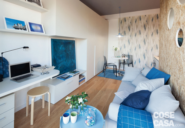 zona giorno del monolocale, divano in tessuto chiaro, cuscini blu, parete in OSB con oblò, zona tv, armadio a ponte in laccato bianco, postazione studio, sgabello in legno, lampada da tavolo orientabile zona giorno del monolocale, divano in tessuto chiaro, cuscini blu, parete in OSB con oblò, zona tv, armadio a ponte in laccato bianco, postazione studio, sgabello in legno, lampada da tavolo orientabile