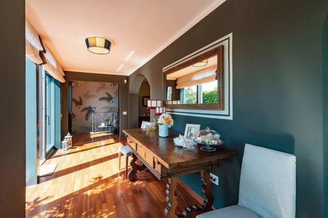 Casa ristrutturata in stile classico moderno, corridoio verandato con ampia scrivania in legno scuro e specchiera. Pavimento in parquet, pareti grigie e arco.: Casa ristrutturata in stile classico moderno, corridoio verandato con ampia scrivania in legno scuro e specchiera. Pavimento in parquet, pareti grigie e arco.: