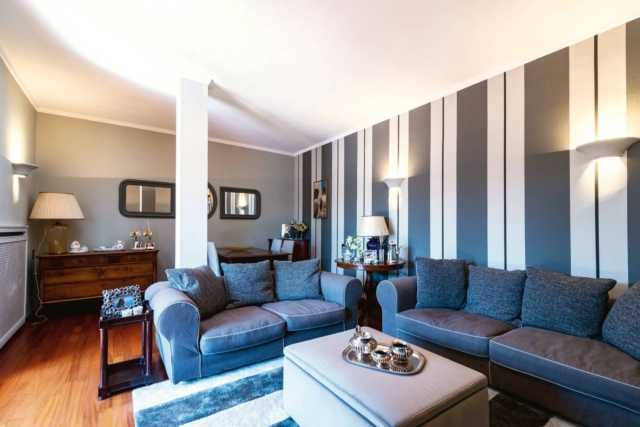 Soggiorno casa ristrutturata in stile classico moderno, divani grigi dalle forme morbide con ampi braccioli, pareti a strisce bianche e grigie Soggiorno casa ristrutturata in stile classico moderno, divani grigi dalle forme morbide con ampi braccioli, pareti a strisce bianche e grigie