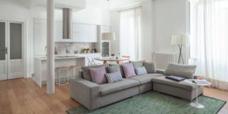 open space del bilocale moderno, divano con chaise longue in tessuto grigio, tappeto, portefinestre, pilastro, cucina, parquet