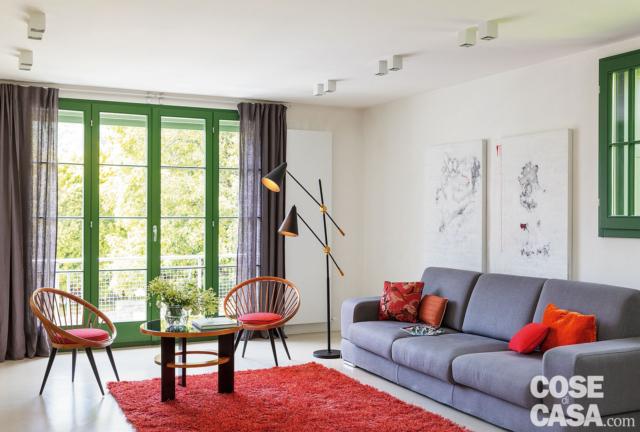 soggiorno della mini casa, divano grigio, cuscini e tappeto rossi, portafinestra, tende, lampada da terra, sedie tonde rosse, pavimento in resina, faretti a soffitto soggiorno della mini casa, divano grigio, cuscini e tappeto rossi, portafinestra, tende, lampada da terra, sedie tonde rosse, pavimento in resina, faretti a soffitto