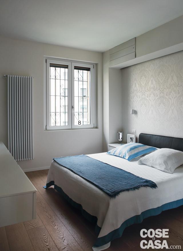 camera matrimoniale, letto, plaid blu, parete rivestita con carta da parati, rientranza, finestra con inferriate, radiatore d'arredo, parquet a doghe, comodino in laccato bianco camera matrimoniale, letto, plaid blu, parete rivestita con carta da parati, rientranza, finestra con inferriate, radiatore d'arredo, parquet a doghe, comodino in laccato bianco
