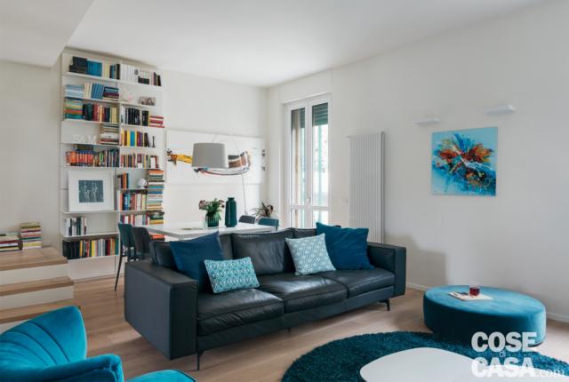 soggiorno della casa su due livelli, zona conversazione, divano in pelle, cuscini azzurri, pouf tondo, poltroncina avvolgente, tappeto, libreria a giorno, portafinestra soggiorno della casa su due livelli, zona conversazione, divano in pelle, cuscini azzurri, pouf tondo, poltroncina avvolgente, tappeto, libreria a giorno, portafinestra