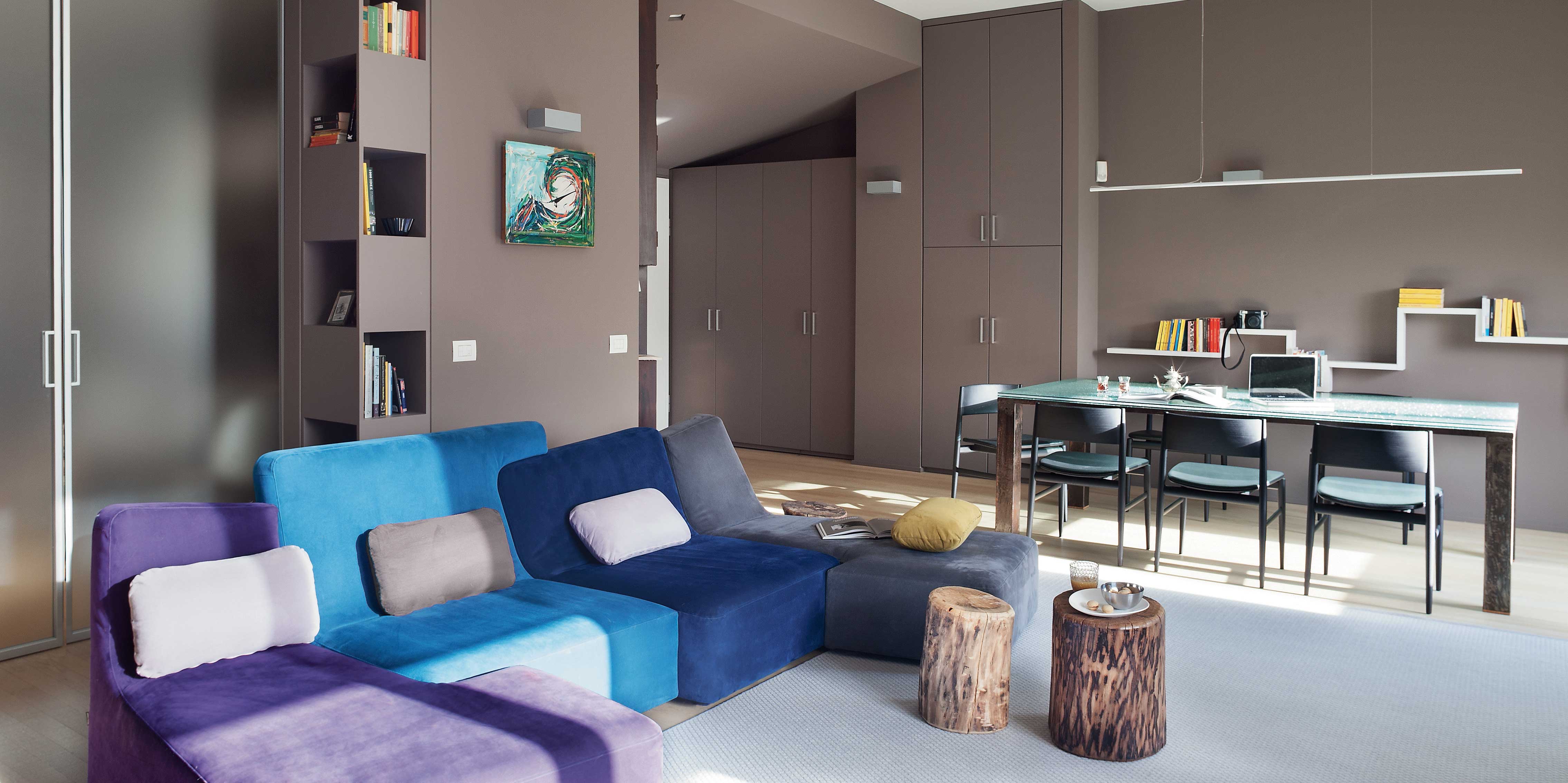 soggiorno, pareti grigio tortora, porta vetrata, divano destrutturato azzurro e viola, cuscini, tavolini a forma di tronco, tavolo da pranzo con piano in vetro, libreria a colonna, ripiani, tappeto grigio
