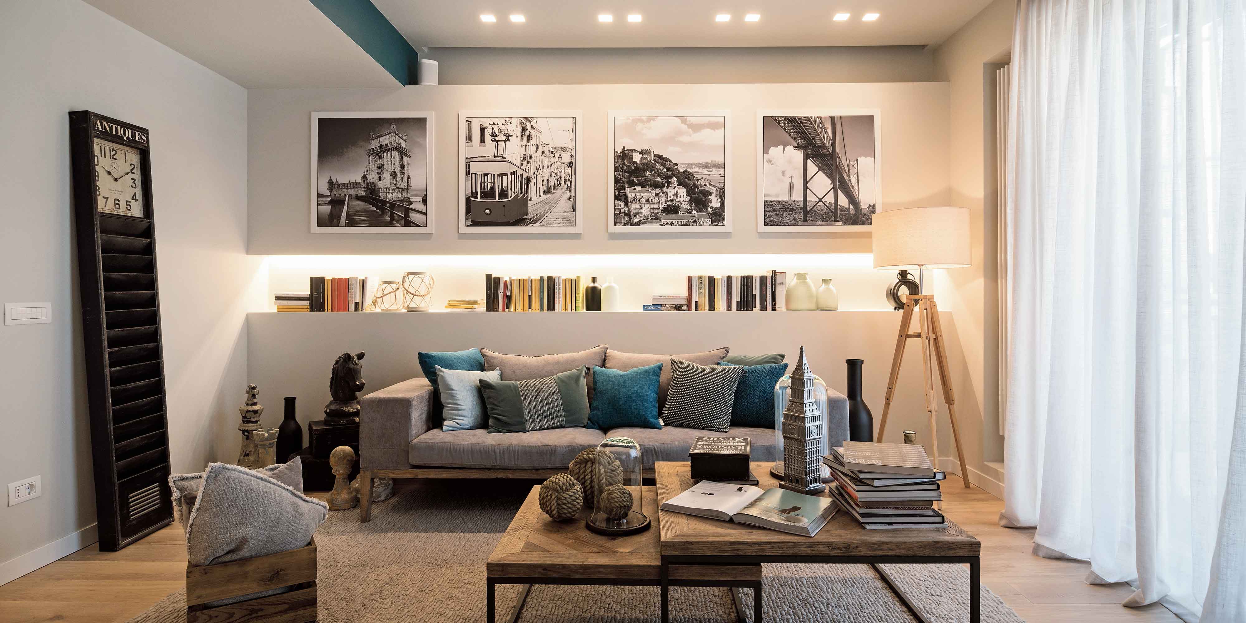 soggiorno aperto, tendaggi bianchi, faretti incassati, controparete luminosa, divano grigio, lampada da terra con treppiede, coffee table, soprammobili, pouf, bocchette impianto canalizzato, tappeto, stampe incorniciate