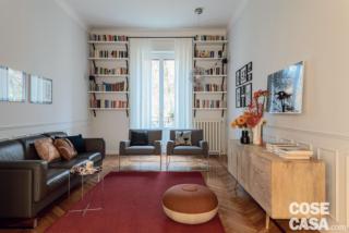 In soggiorno i ripiani della libreria - realizzata su disegno da Giacomo Moor - incorniciano il rettangolo della finestra; le pareti perimetrali sono decorate nella fascia bassa con profili in gesso effetto boiserie. Il divano rivestito in pelle è Cab di Truedesign, i cuscini sono foderati con tessuti Dedar. Il tavolino rotondo cromato è Habibi di e15, le due poltrone sono le Charles Armchairs di Moooi; stessa produzione per la lampada a sospensione sferica modello Raimond. Sulla madia in legno di Maisons du Monde, vasi e candeliere sono in vendita presso lo store Fritz Hansen; quello bianco con i rami di nandina è Bambu di Danese, design Enzo Mari. Il pouf tondo Large di Celice Manz è in vendita da Fritz Hansen. Il tappeto in lana è Poppy di Kasthall.