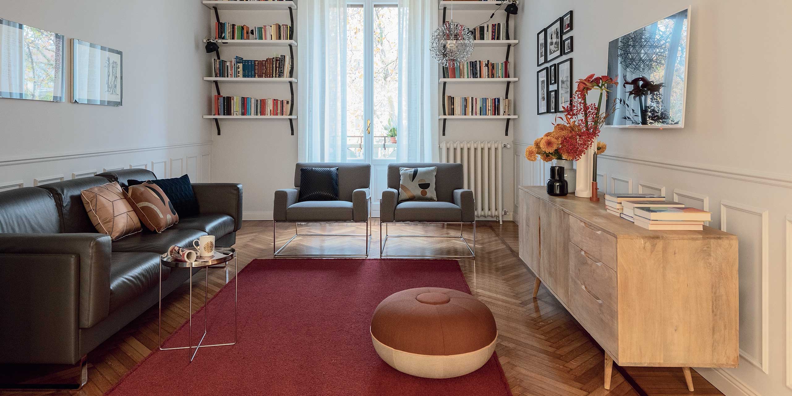 soggiorno del trilocale, finestra, libreria a portale, divano in pelle, poltroncine, madia in legno, pouf tondo, tappeto rosso, parquet a spina di pesce, radiatore in ghisa, quadri