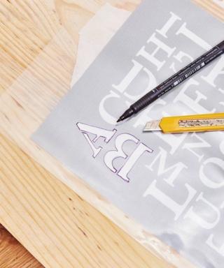 Recuperare dal web le sagome di lettere e numeri per stencil e stamparle secondo il formato desiderato, variando però le misure. Appoggiare sopra le lettere il foglio di acetato e, con il pennarello nero indelebile a punta sottile, ricalcare i profili. Trasferire il foglio sopra una superficie d'incisione, per esempio una base in legno di riciclo oppure un tappetino da taglio. Con il cutter, incidere con precisione le lettere, in modo da ricavare la/le mascherine.