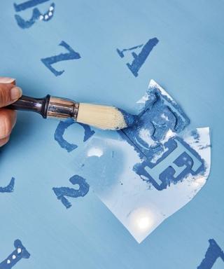 Spruzzare un velo di colla spray riposizionabile sul retro delle mascherine; eliminare l’eccedenza premendola sulla carta da pacco. Disporle sul piano e, con il pennello a punta tonda e la pittura bluette (tamponare il di più con un foglio), picchiettare delicatamente sulla parte incisa fino a riempire l'intera lettera. A lavoro ultimato rimuovere la sagoma e fare asciugare. Proseguire così, alternando lettere e numeri. Impreziosire con tocchi di bianco.