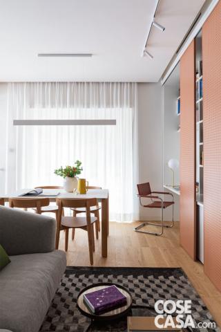 soggiorno del trilocale, zona pranzo, tavolo, sedie, vaso bianco, vetrata, tendaggio, lampada a sospensione lineare, controsoffitto, postazione studio, sedia, faretti orientabili a soffitto, libreria con pannelli forati scorrevoli, divano grigio, tavolino, tappeto