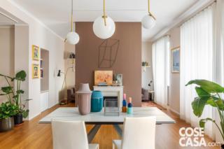 open space dell'appartamento ristrutturato, zona pranzo, tavolo con piano bianco, sedie, lampade a sospensione di design, vasi, portefinestre, tendaggi bianchi, quinta tinteggiata color tortora, camino a bioetanolo, parquet di bambù