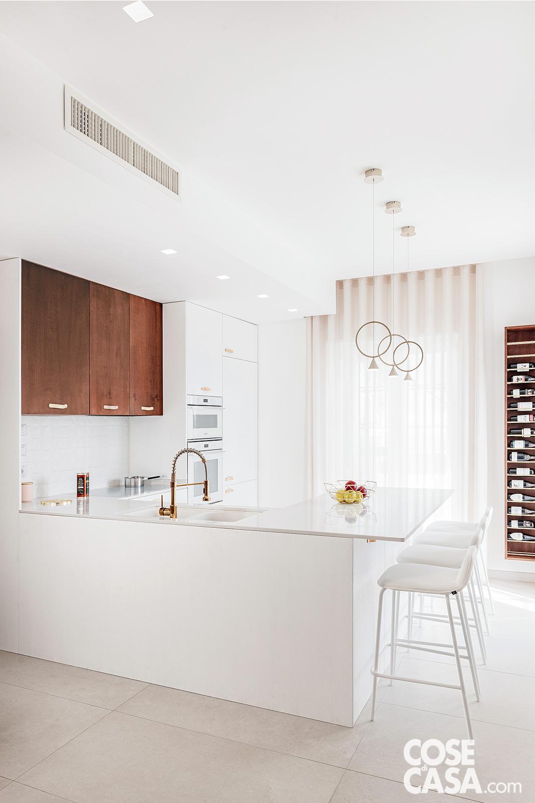 Bancone cucina bianco a ferro di cavallo, con sedie bianche, e pensili legno scuro. A destra, dettaglio di una cantinetta a muro con ripiani per bottiglie di vino