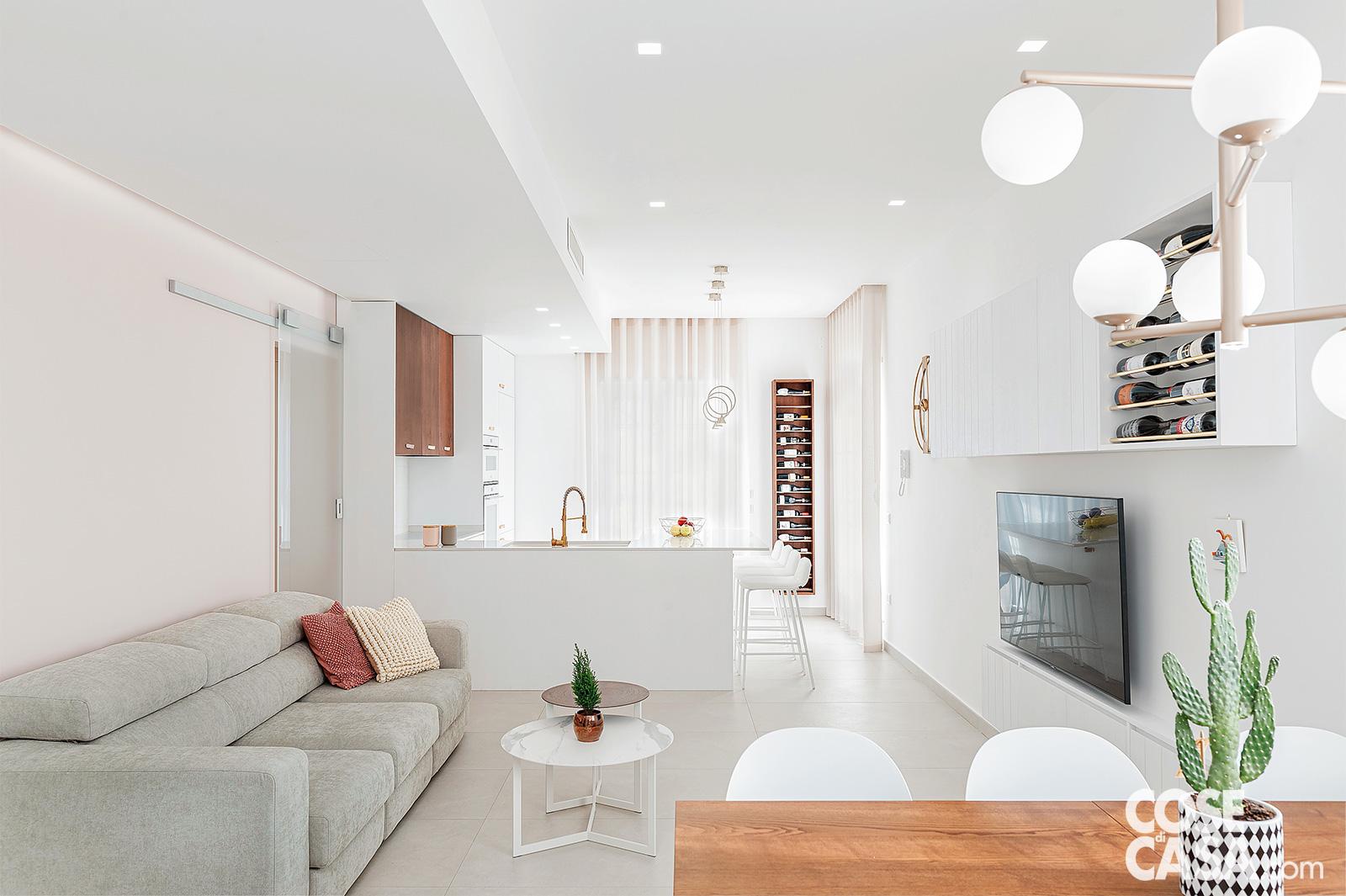 Living open space con cucina a vista, tavolo da pranzo in essenza e divano grigio. Dettagli di design moderno con arredi bianchi e tocchi in legno.