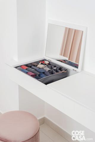 Scrivania bianca con specchio e contenitore integrato, con divisori per accessori come cravatte e orologi. Sgabello tondo rosa di fronte alla scrivania.