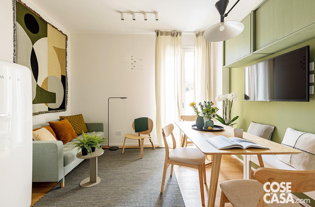 Il bilocale si apre sulla zona giorno che integra living, pranzo e angolo cottura. Il divano appoggiato alla parete, rivestito in tessuto grigio, è Landskrona di Ikea; i cuscini sono di Moroso e di Christian Fischbacher. Arazzo a parete Entire di Ferm Living. La scultura metallica a sinistra della portafinestra è di Mobili Futura. Il tavolino rotondo è di Moroso, il tappeto di Kasthall, il plaid appoggiato sullo schienale della sedia di Christian Fischbacher. Nella zona pranzo il tavolo rettangolare in legno è il modello Keira, le sedie sono le Cloda, tutto di Sklum. Sul piano, mug bianca di Le Creuset e vasi di Zara Home e Asa Selection. Le panchette bianche appoggiate alla parete sono le Ekedalen di Ikea con cuscini di Zara Home.