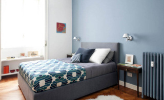 Tavolino vintage in legno, accostato a un letto tessile grigio, diventa comodino. Tavolino vintage in legno, accostato a un letto tessile grigio, diventa comodino.