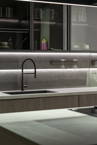 La zona lavaggio è servita da uno schienale magnetico e luminoso Duetto in Laminam® Pietra Grey, lo stesso materiale usato per i top. È attrezzato con mensole, portacalici e porta spezie della collezione di accessori magnetizzati Origami. Sopra, vetrine Vogue composte da vetro fumé, schiena in laminato riga termocotto, telaio nero montato con cerniera a scomparsa e luci interne.