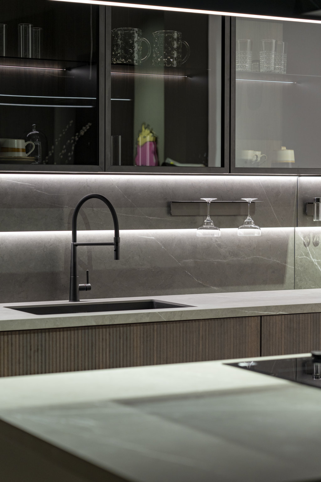 La zona lavaggio è servita da uno schienale magnetico e luminoso Duetto in Laminam® Pietra Grey, lo stesso materiale usato per i top. È attrezzato con mensole, portacalici e porta spezie della collezione di accessori magnetizzati Origami. Sopra, vetrine Vogue composte da vetro fumé, schiena in laminato riga termocotto, telaio nero montato con cerniera a scomparsa e luci interne.