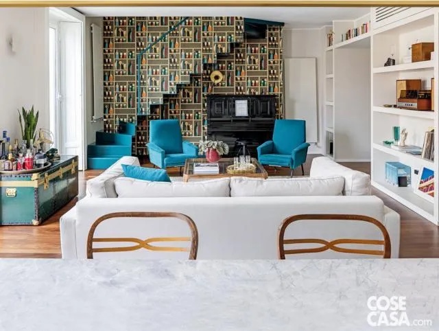 Nella zona giorno la carta da parati (Cocktails coll. Fornasetti di Cole & Son) che riveste a tutt’altezza la parete modifica visivamente la profondità del volume. Nel motivo trompe l’oeil del decoro – un bar con scaffali e bottiglie – si mimetizza la sagoma azzurra della rampa a giorno in lamiera piegata e resinata che sale in linea retta. Sotto la scala trova posto il pianoforte. Le poltroncine rivestite in tessuto azzurro sono di Mediatrade Casa d’Aste, il tavolino in legno con piano in vetro è di Fratelli Levaggi. A sinistra, il baule verde utilizzato come mobile bar è un pezzo di famiglia d’antiquariato. Nella zona giorno la carta da parati (Cocktails coll. Fornasetti di Cole & Son) che riveste a tutt’altezza la parete modifica visivamente la profondità del volume. Nel motivo trompe l’oeil del decoro – un bar con scaffali e bottiglie – si mimetizza la sagoma azzurra della rampa a giorno in lamiera piegata e resinata che sale in linea retta. Sotto la scala trova posto il pianoforte. Le poltroncine rivestite in tessuto azzurro sono di Mediatrade Casa d’Aste, il tavolino in legno con piano in vetro è di Fratelli Levaggi. A sinistra, il baule verde utilizzato come mobile bar è un pezzo di famiglia d’antiquariato.