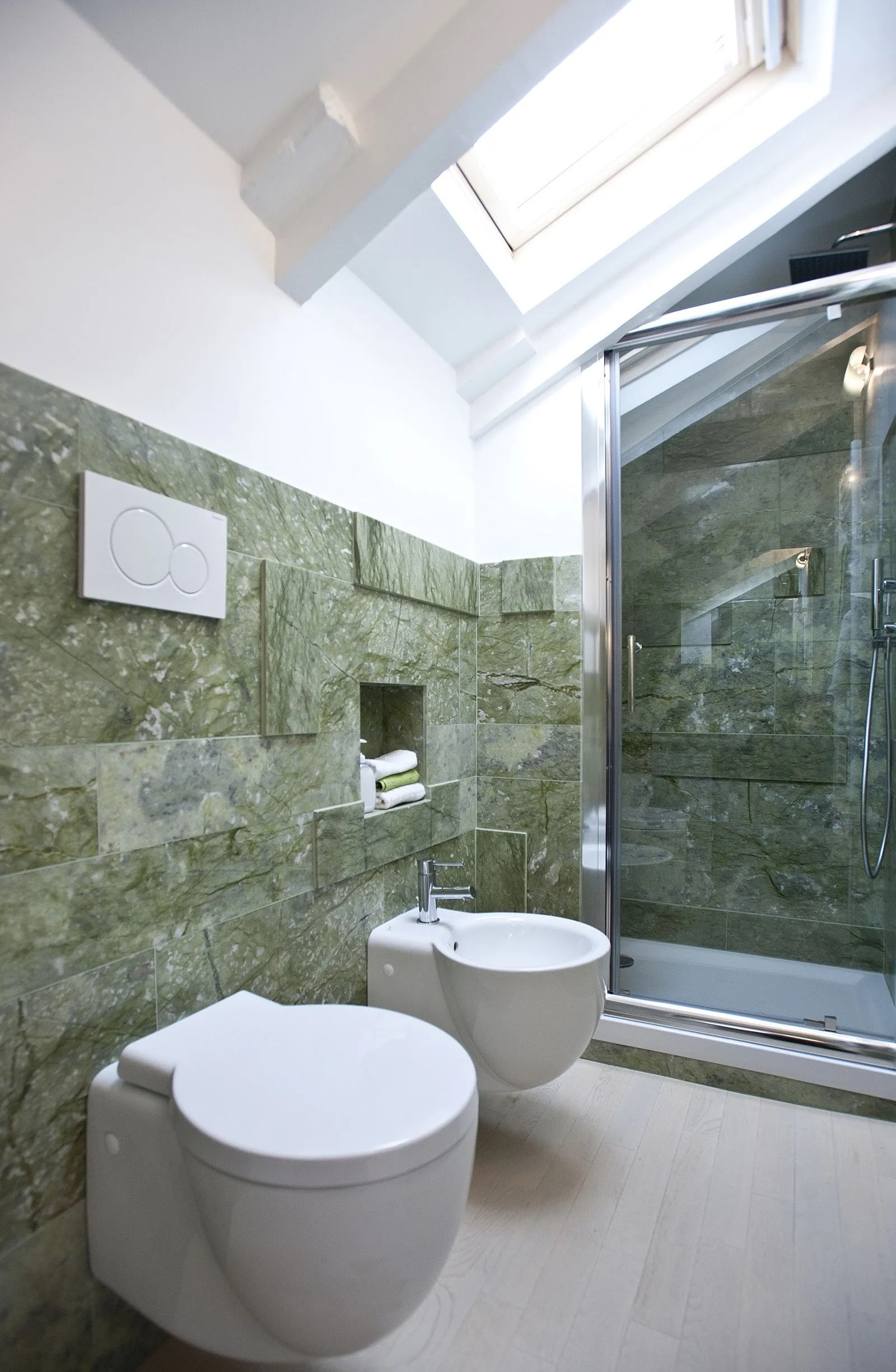 Nel piccolo bagno sottotetto, i sanitari sospesi (di Azzurra) e il box doccia di cristallo trasparente su misura esaltano il senso di leggerezza, controbilanciato dall’elegante gioco materico del rivestimento di marmo verde Ming, con lastre di spessore e dimensioni diverse, abbinato al pavimento in legno. Foto: Jule Hering. Progetto: arch. Fabrizio Arcoini e arch. Martina Margaria – Studio Abita. Nel piccolo bagno sottotetto, i sanitari sospesi (di Azzurra) e il box doccia di cristallo trasparente su misura esaltano il senso di leggerezza, controbilanciato dall’elegante gioco materico del rivestimento di marmo verde Ming, con lastre di spessore e dimensioni diverse, abbinato al pavimento in legno. Foto: Jule Hering. Progetto: arch. Fabrizio Arcoini e arch. Martina Margaria – Studio Abita.