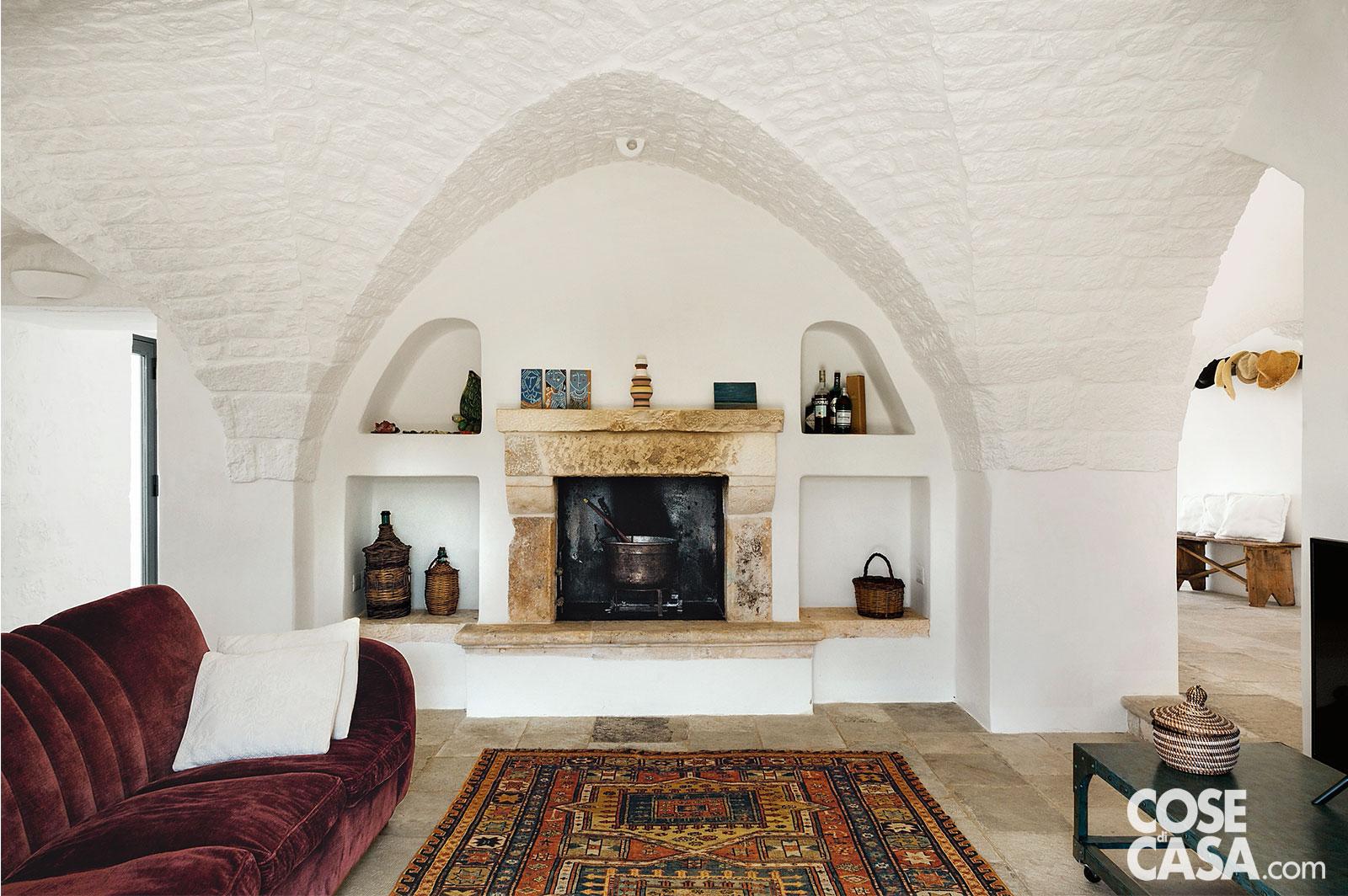 Il camino al centro del salottino ricavato nell’arco di un trullo, tinto di un bianco candido, esalta la pietra locale del pavimento. Il focolare si prende la scena in questa zona giorno in cui spiccano un divano in velluto e il tappeto persiano in palette - Foto: Studio Roy. Progetto: arch. Azzurra Garzone.