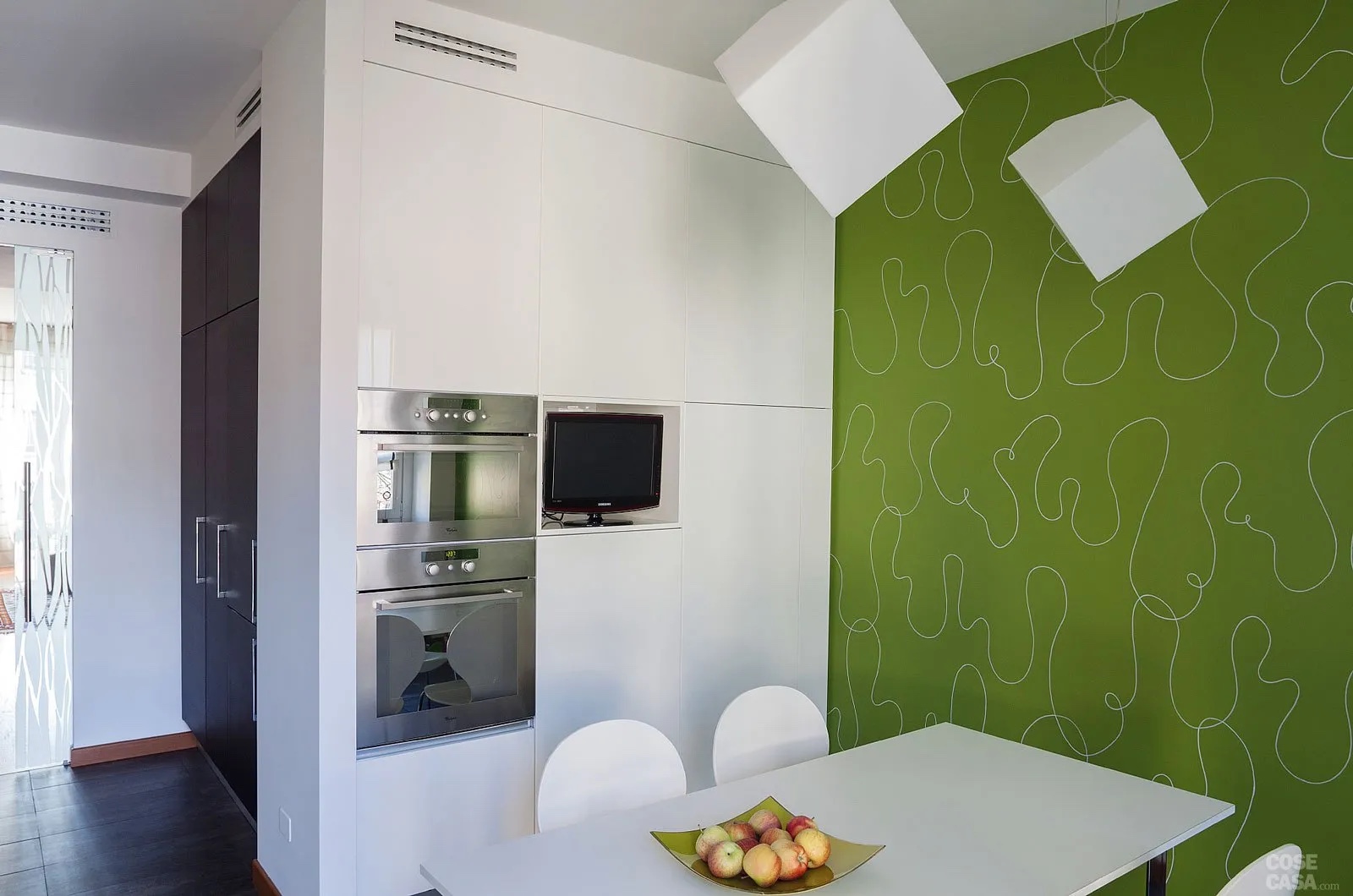 Dietro un'armadiatura in laccato bianco, che integra vani contenitori e forno, si apre l'area pranzo arredata con tavolo e sedie anch'esse bianche: qui a sorpresa, la parete laterale è rivestita con una tappezzeria verde a decori bianchi, che spicca e crea un contrasto sia per il colore molto forte sia per il disegno a linee ondulate che spezza visivamente il rigore e la linearità del resto. La zona pranzo, organizzata in un angolo della cucina, è evidenziata dalla tappezzeria verde a decori bianchi della parete laterale.