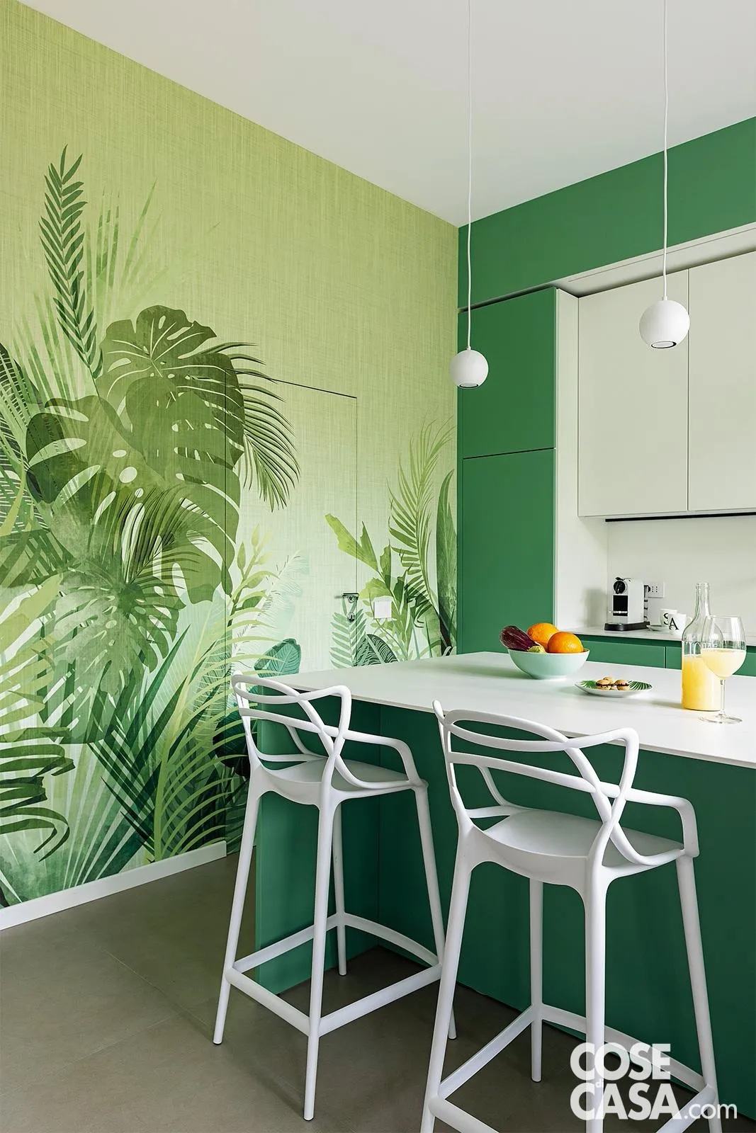 Tappezzeria jungle in cucina che mimetizza la porta filomuro. Gli sgabelli da bar bianchi accostati al bancone snack dell’isola sono il modello Masters di Kartell, design Philippe Starck. Le lampade a sospensione sferiche sono di Ideal Lux. La carta da parati a motivo jungle sulla parete a sinistra è di Inkiostro Bianco. Foto: Cristina Galliena Bohman, styling Laura Mauceri. Progetto: arch. Francesca Bonavia, Studio MB5 Architettura.