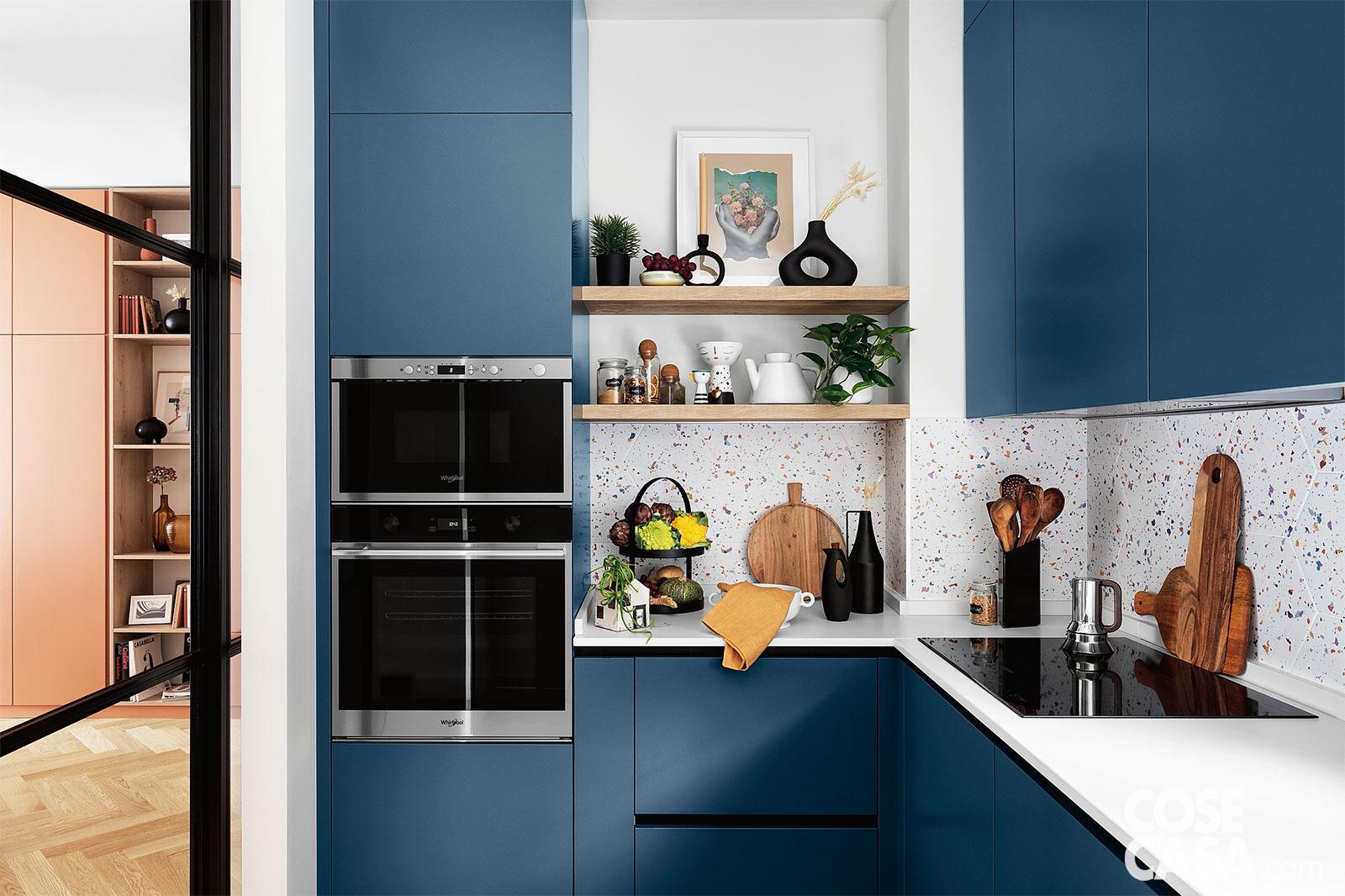 Una cucina ad angolo dalle linee pulite e semplici con pannelli in blu navy a contrasto con il piano di lavoro bianco, l’alzata puntinata ed elementi in legno naturale e neri a contrasto. Il risultato è delicatamente vintage.