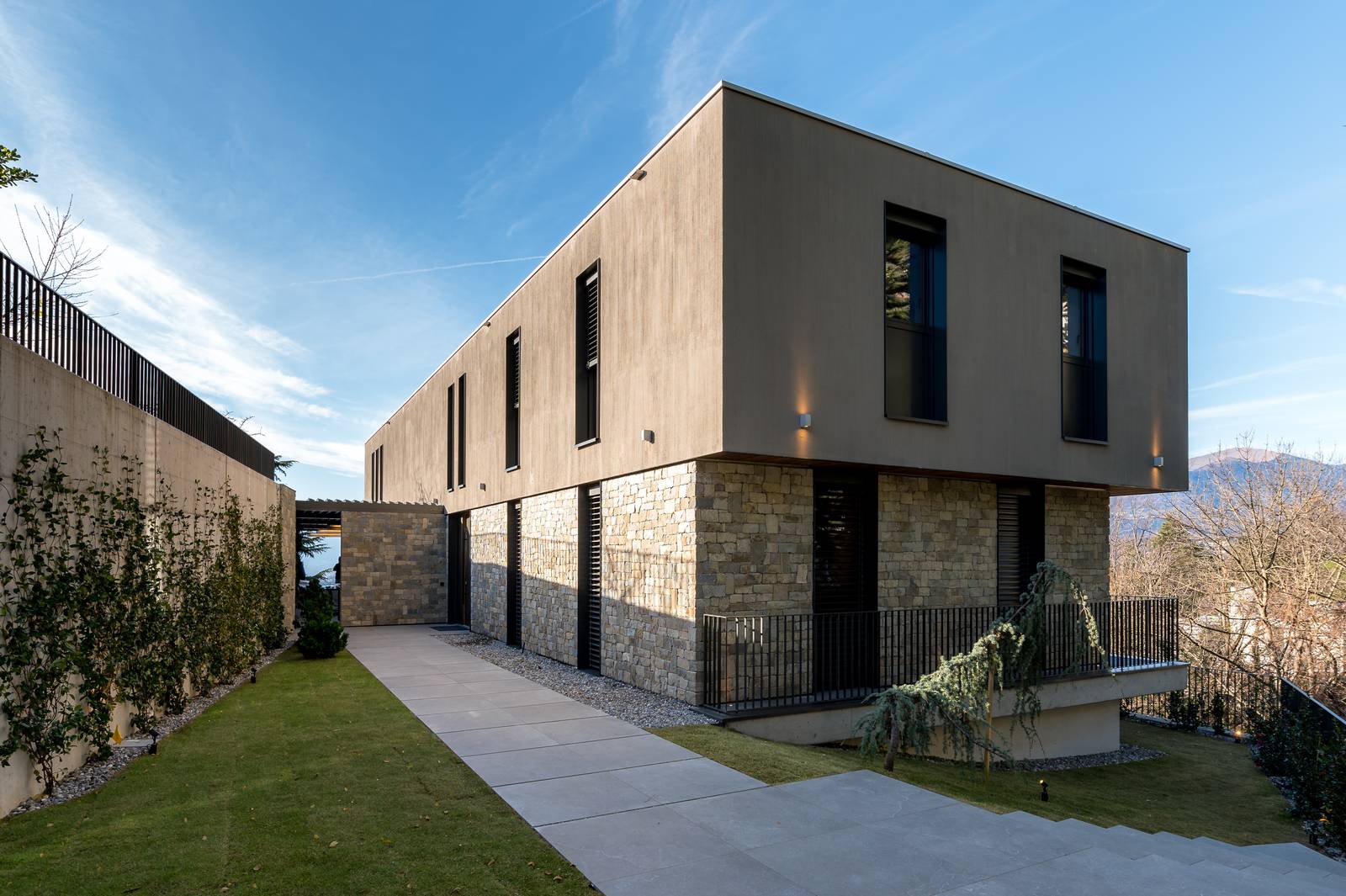 Vista esterna della villa, che mostra il volume essenziale e scultoreo della struttura. Si evidenziano le pareti in pietra a vista al piano terra e la facciata superiore liscia. Il camminamento esterno è sempre in Lithos Desert, ma con spessore superiore, di 20 mm. Vista esterna della villa, che mostra il volume essenziale e scultoreo della struttura. Si evidenziano le pareti in pietra a vista al piano terra e la facciata superiore liscia. Il camminamento esterno è sempre in Lithos Desert, ma con spessore superiore, di 20 mm.