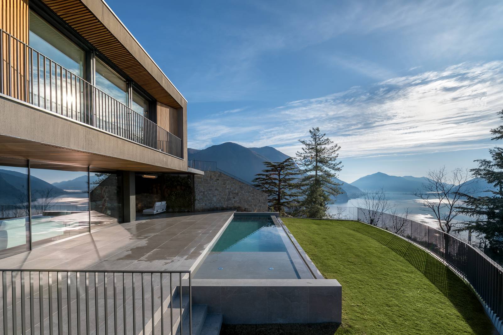 L'area della piscina a sfioro, con vista sul lago e le montagne. Il bordo piscina è rivestito da piastrelle (Lithos Desert 20 mm) con finitura bocciardata, garantendo continuità estetica ma anche sicurezza antiscivolo. L'area della piscina a sfioro, con vista sul lago e le montagne. Il bordo piscina è rivestito da piastrelle (Lithos Desert 20 mm) con finitura bocciardata, garantendo continuità estetica ma anche sicurezza antiscivolo.