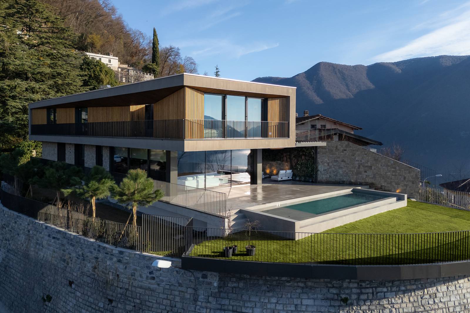 Veduta aerea che mostra la villa inserita nel contesto paesaggistico, con la sua architettura moderna e la presenza della piscina e degli ampi spazi esterni. Si apprezzano le facciate con elementi in legno e pietra. Veduta aerea che mostra la villa inserita nel contesto paesaggistico, con la sua architettura moderna e la presenza della piscina e degli ampi spazi esterni. Si apprezzano le facciate con elementi in legno e pietra.