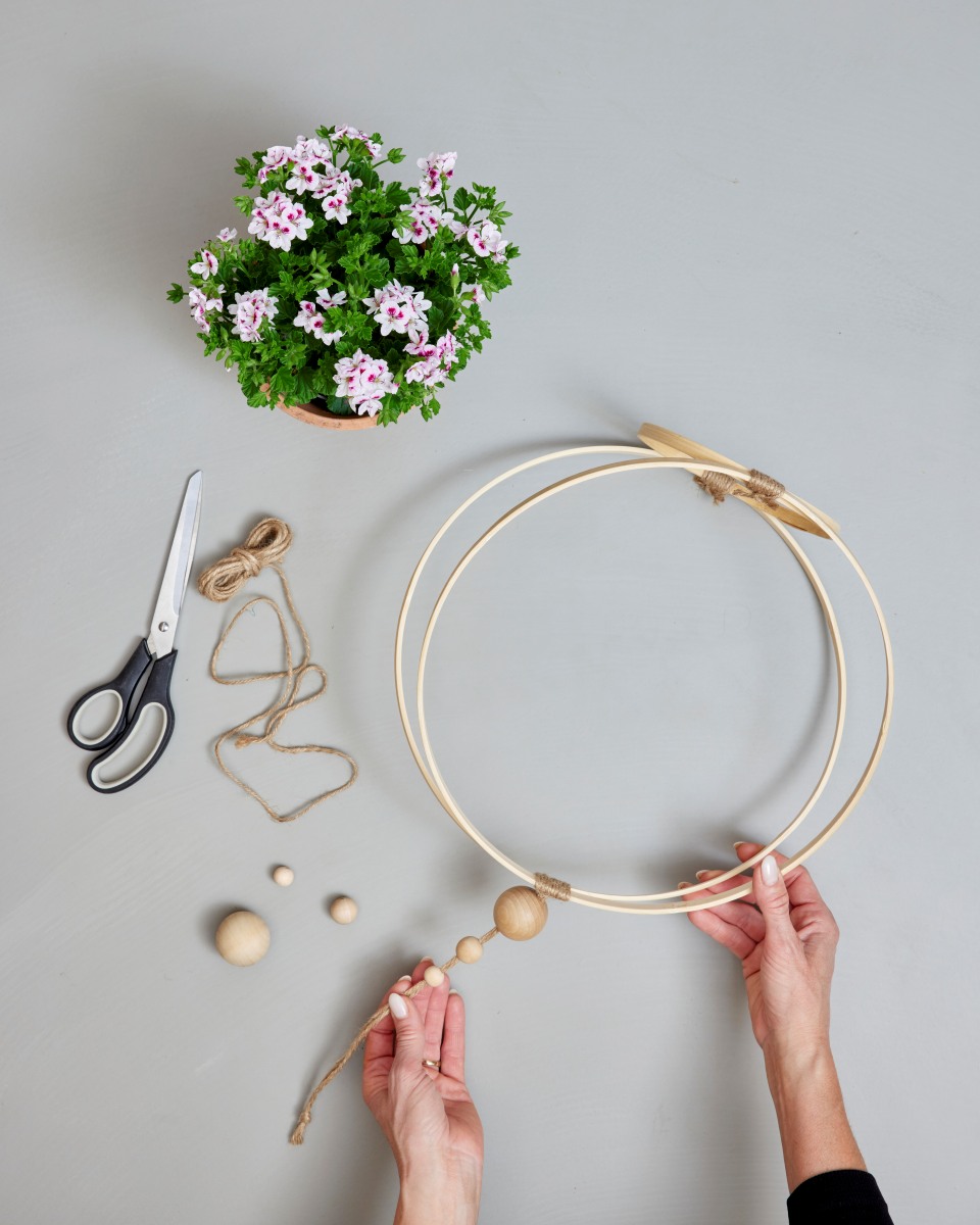 È il momento di creare il sistema di sospensione del vaso infilando le perline di legno sullo spago in ordine crescente, facendo in modo che la più grande si appoggi sugli anelli. Dopo aver fissato saldamente lo spago nel punto in cui i due cerchi più grandi si toccano, si può avvolgere lo spago intorno al nastro adesivo per nasconderlo e ottenere un aspetto più elegante. Si può infine infilare il vaso di terracotta nel cerchio più piccolo e appenderlo dove si desidera. Copyright immagine Pelargonium for Europe