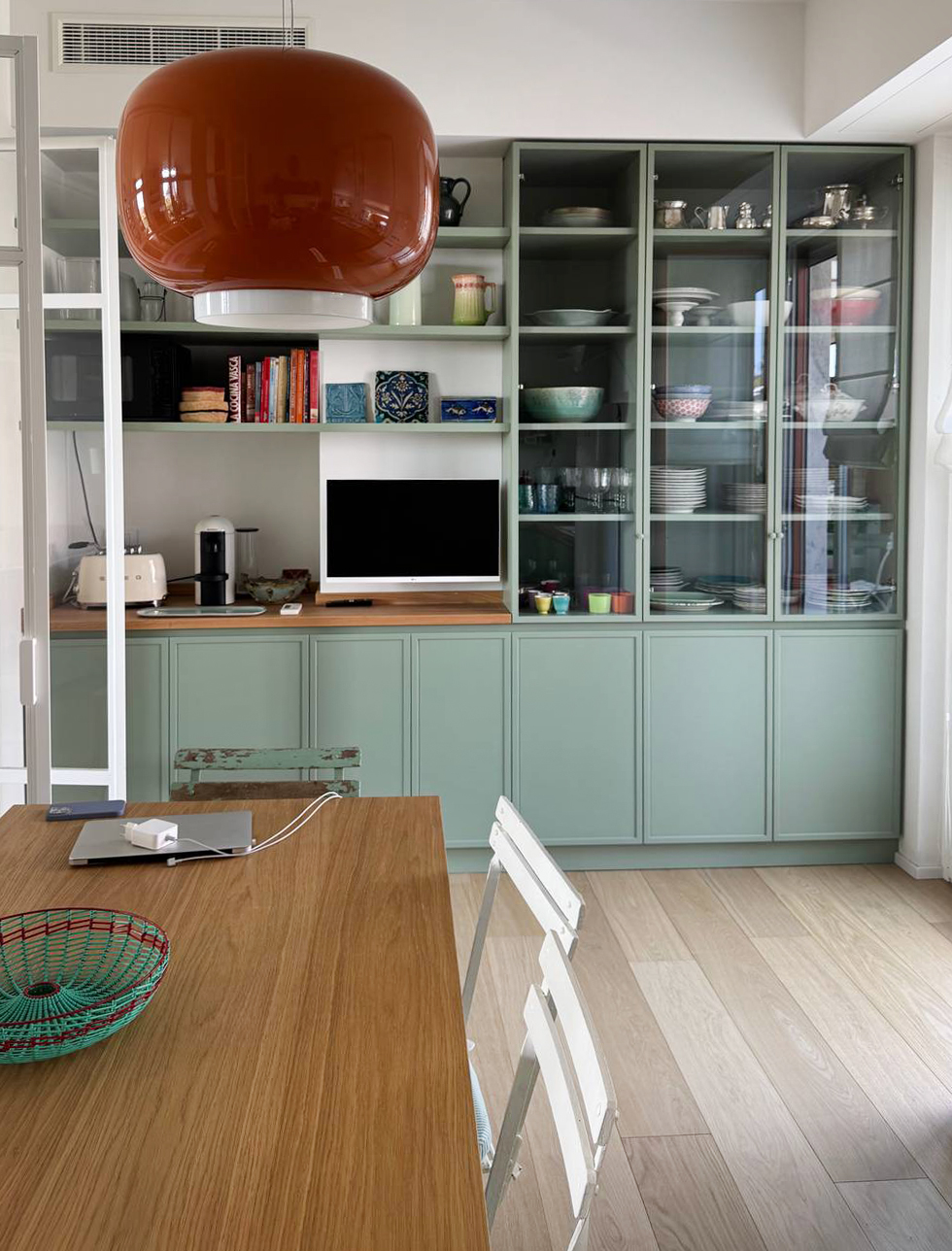 Un verde di media intensità, sobrio ma distintivo, veste tutti i mobili di questa cucina, inclusi quelli a vetrina, creando un insieme coerente e caldo. Le pareti bianche amplificano la luminosità e lasciano emergere il dialogo tra colore e materia, mentre il pavimento in rovere chiaro riprende il tono del piano di lavoro, rafforzando il senso di continuità e naturalezza.