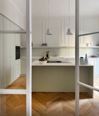 L’apparente semplicità del bianco viene reinterpretata in chiave decorativa. La parete è scandita da quattro ampie cornici in gesso, dal sapore classico e misurato, che incorniciano la composizione e fungono da quinta scenica per i mobili color verde pistacchio chiarissimo. Una lunga mensola orizzontale, spessa 6 cm, attraversa l’intera parete, segnando una linea grafica netta e ordinatrice. Il risultato è uno spazio luminoso, fresco e bilanciato tra tradizione e modernità.