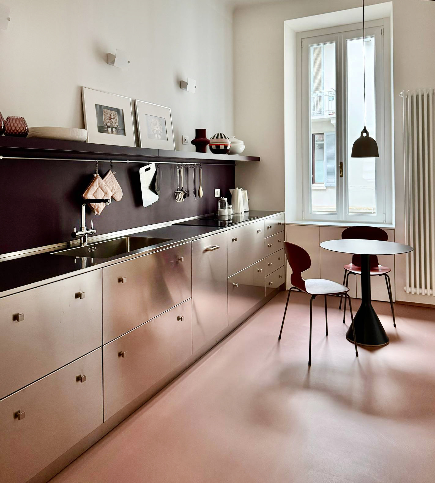 I mobili in acciaio - come quelli delle cucine industriali dei ristoranti - possono apparire troppo freddi. Ma ecco un esempio che dimostra come anche un materiale tecnico possa cambiare completamente a seconda di come viene abbinato: il progetto che sovverte la percezione dell’acciaio, trasformandolo da materiale tecnico a protagonista di uno spazio caldo e accogliente. Come? Grazie ai colori caldi scelti per pavimento (in resina color terracotta che diffonde una luce morbida che si riflette sulle superfici metalliche, ammorbidendole) e backsplash (in resina color melanzana, che introduce una nota profonda e avvolgente). L’atmosfera è domestica e sofisticata, lontana dal rigore delle cucine professionali, un effetto accentuato anche dall’utilizzo di pomoli dal sapore retrò per aprire le ante.