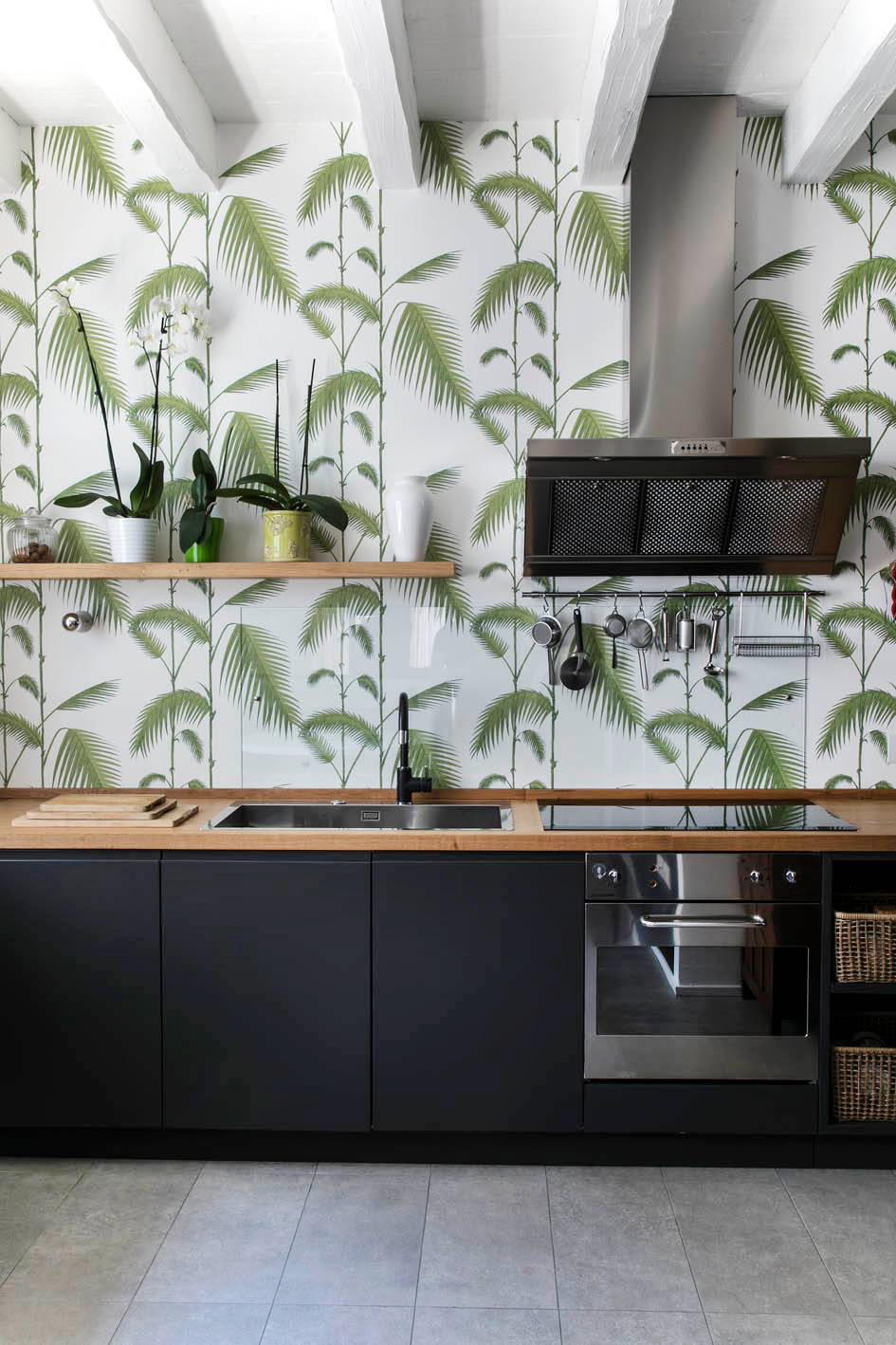 Pensata per chi desidera che la parete di fondo diventi l’elemento focale della cucina, questa soluzione affida alla carta da parati il compito di definire l’identità dell’ambiente. Rivestirla con un motivo decorativo è una scelta di grande impatto visivo, capace di trasformare radicalmente la percezione dello spazio. In questo progetto, il disegno botanico verde su fondo bianco cattura immediatamente l’attenzione e conferisce un carattere scenografico all’insieme, bilanciato dalla sobrietà dei mobili neri, dalle superfici lisce e dalle linee essenziali. Se la parete fosse lasciata bianca, l’effetto complessivo cambierebbe completamente: da decorativo e ricco di personalità diventerebbe minimale e rarefatto.