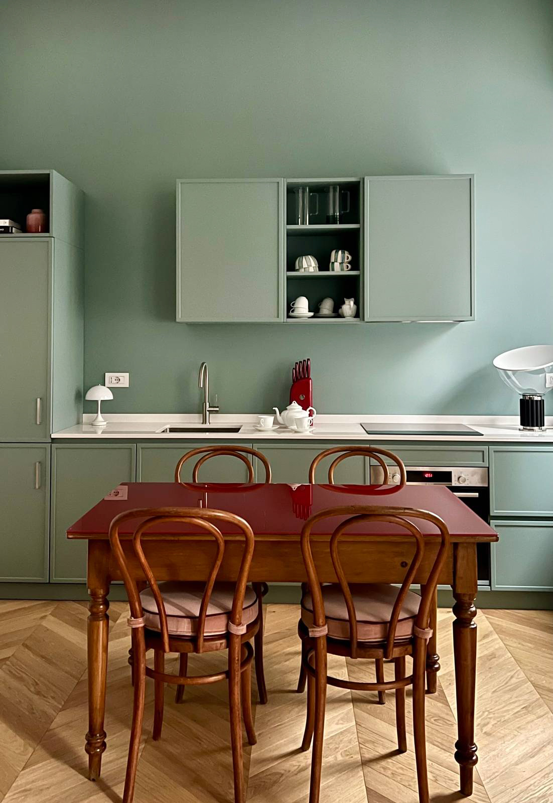 Una scelta progettuale netta e contemporanea: parete e mobili sono trattati con lo stesso tono di azzurro-verde profondo, generando un effetto “a scatola” o Color Block, di grande impatto visivo. L’uniformità cromatica avvolge lo spazio e lo rende coerente e immersivo, mentre la disposizione dei pensili – centrati sulla parete e non a tutta larghezza – introduce un delicato gioco di pieni e vuoti che aggiunge dinamismo e profondità.